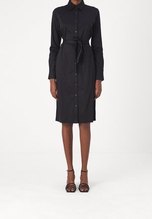 Vrouw die een zwarte blousejurk met knopen tot op de knie draagt, met een ceintuur in de taille en zwarte sandalen met hoge hakken en bandjes, staand tegen een witte achtergrond.