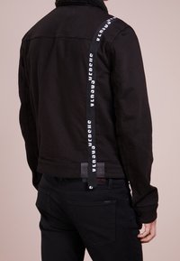 Giacca di denim nera con colletto in sherpa, caratterizzata da una fascia bianca con logo e un'etichetta con marchio all'orlo. Paio di pantaloni neri visibili sotto.