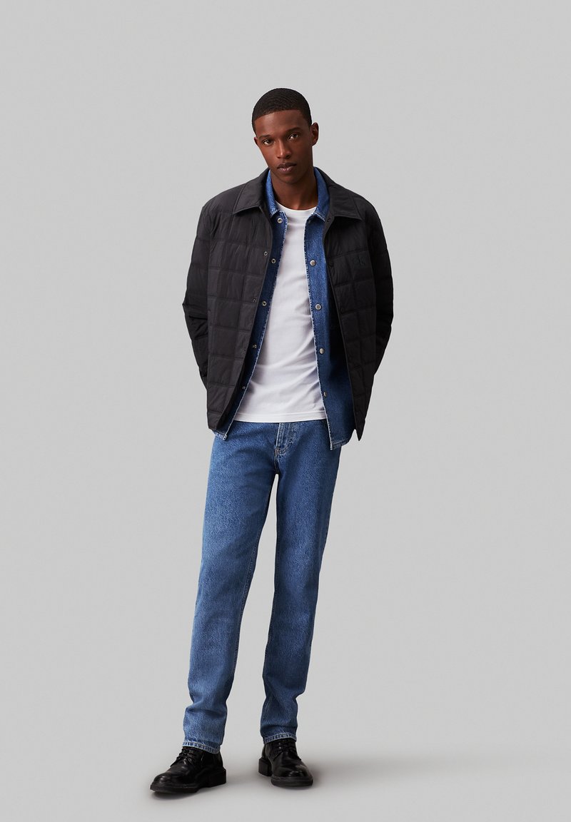 Jachetă neagră matlasată peste o cămașă din denim, un tricou alb și blugi. Pantofi negri completează aspectul. Fundalul este gri simplu.