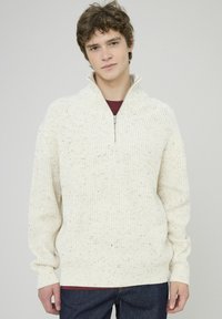Pull en tricot écru avec un col montant zippé à mi-hauteur, présentant un motif texturé et des éclats de couleur partout, porté sur une chemise bordeaux.