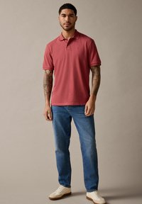Camisa polo de algodón roja con mangas cortas, combinada con jeans de mezclilla azules y zapatillas beige. El modelo se encuentra de pie contra un fondo neutro.