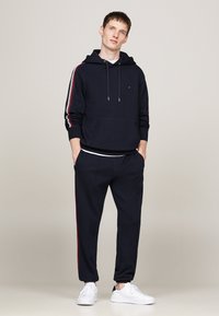 Sudadera azul marino con rayas laterales rojas y blancas, detalle de bolsillo y cordón. Combinada con joggers azul marino a juego y zapatillas blancas.
