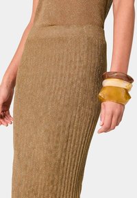 Strickkleid aus Rippstrick in Bronze mit taillierter Form. Das Handgelenk geschmückt mit klobigen, durchsichtigen Harzarmreifen in Bernstein- und Creme-Tönen.
