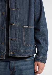 Giacca di jeans di colore blu scuro con vestibilità rilassata, dotata di bottoni in ottone, cuciture a contrasto e una tasca applicata con dettaglio del marchio.