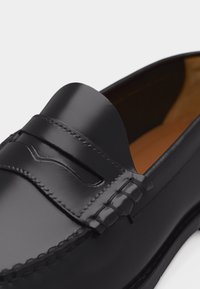 Μαύρο δερμάτινο παπούτσι τύπου penny loafer με γυαλιστερό φινίρισμα και ραφές σε στυλ μοκασίνι σε λευκό φόντο.