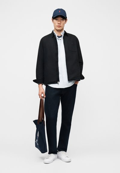 Νέος άνδρας που φοράει ναυτικό καπέλο, μαύρο μπουφάν με φερμουάρ πάνω από ριγέ πουκάμισο, σκούρο παντελόνι, λευκά αθλητικά παπούτσια, κρατώντας ναυτικό τσάντα tote με καφέ λαβές.