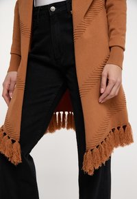 Bruine gebreide cardigan met gestructureerde zigzagpatronen en franjes aan de onderkant, gecombineerd met zwarte high-waisted jeans.