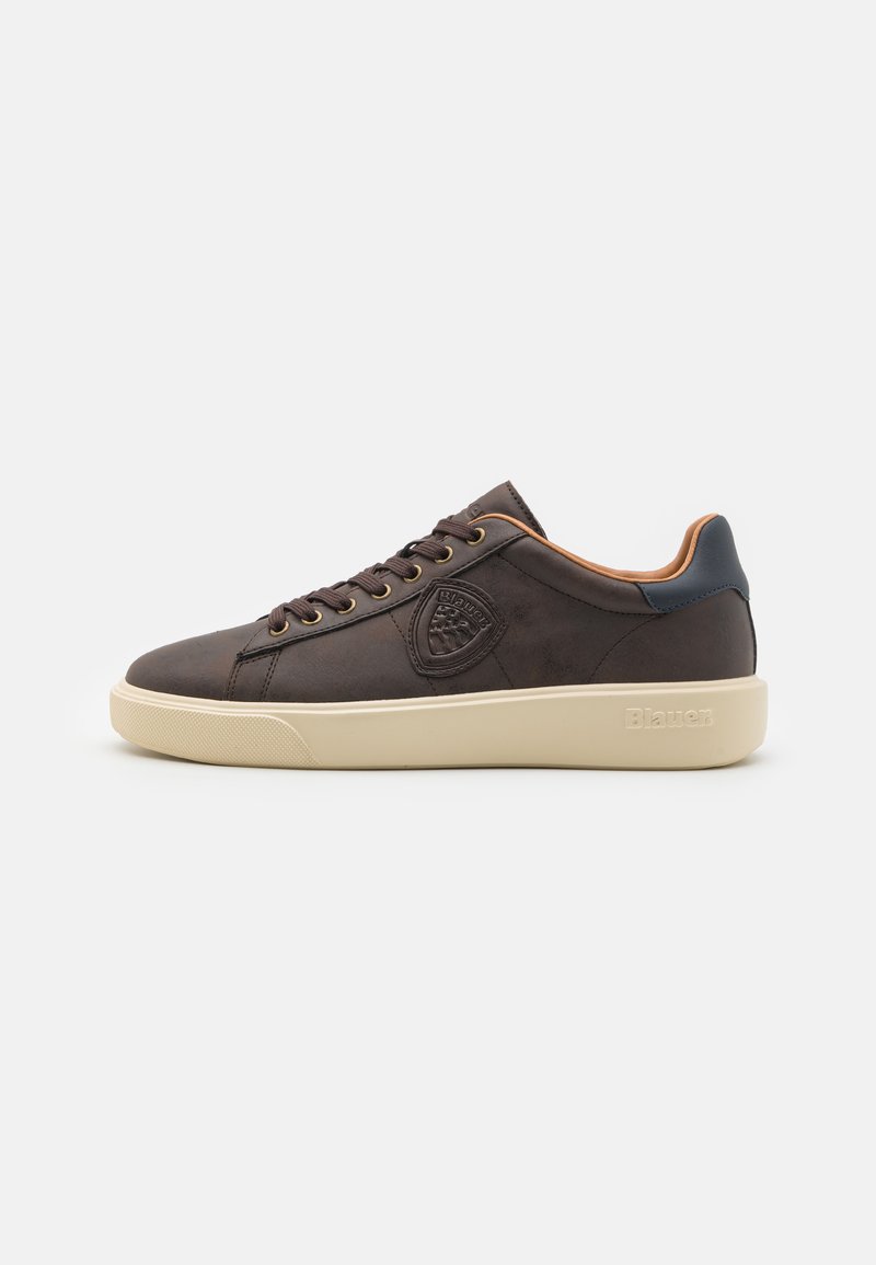 Zapatilla de cuero marrón con suela de goma crema, punta redonda y ojales dorados. Presenta un parche con el logo en el lateral y un acento en el talón en tono azul marino.