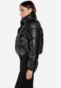 Chaqueta acolchada corta de cuero sintético negro con un alto cuello, cierre frontal y puños elásticos; presenta una textura acolchada y un diseño elegante.
