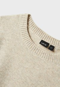 Ljusbeige stickad tröja med ribbad rund halsringning, mjuk textur och taggar som visar storlek och märke.