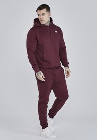 Completo felpa e pantaloni della tuta color bordeaux in un tessuto morbido, con tasca frontale e un piccolo logo sul petto. Indossato con sneakers bianche.