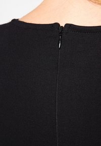 Black, fitted top with a round neckline, featuring a central back seam and a small zipper closure at the nape. Soft, textured fabric.