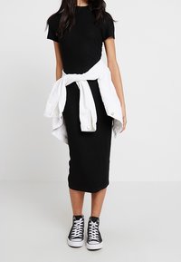 Robe midi noire côtelée à manches courtes, associée à une veste blanche nouée et des sneakers montants noirs. Texture lisse, silhouette ajustée.