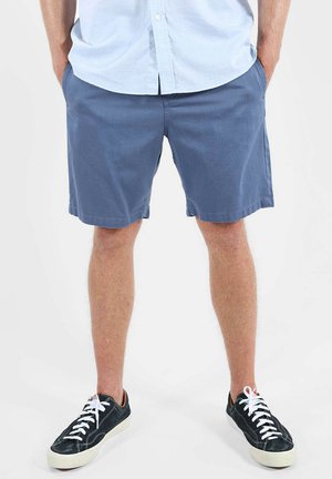 Homme portant un short bleu, une chemise à manches longues bleu clair boutonnée, des baskets noires avec des lacets blancs, debout les mains dans les poches sur fond blanc.
