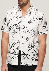 Camisa de manga corta en tela blanca con estampado de bambú negro, presenta un cierre frontal con botones y bolsillos en el pecho, y botones de madera.