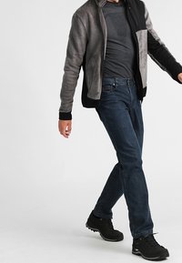 Person som går i en grå och svart jacka, mörkgrå skjorta, blå jeans och svarta vandringsskor framför en enhetlig bakgrund.