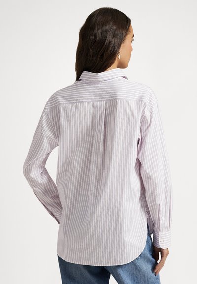 Sieviete ar gariem tumšiem matiem valkā vaļīgu baltu pogājamu kreklu ar plānām vertikālām violetām svītrām un zilas džinsas, raugoties prom.