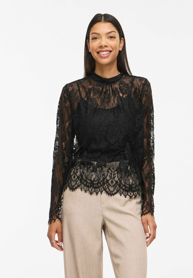 Femme souriante portant un chemisier en dentelle noire à manches longues avec un pantalon beige, debout devant un fond blanc.