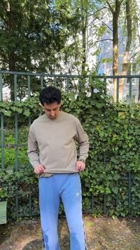 Άνδρας με μπεζ μακρυμάνικο πουκάμισο και ανοιχτό μπλε φόρμα Umbro στέκεται έξω μπροστά σε φράχτη που καλύπτεται από κισσό, κοιτάζοντας προς τα κάτω και κρατώντας το κάτω μέρος του πουκαμίσου.