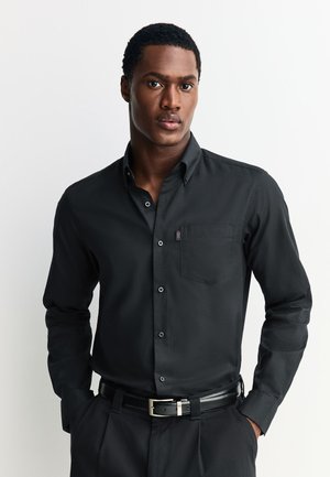 Mies, jolla on musta nappipaita ja mustat housut mustalla vyöllä, seisoo kädet taskuissa yksinkertaisen taustan edessä.