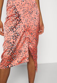 Femme portant une jupe portefeuille corail rose imprimé léopard avec une fente latérale, montrant une partie de ses jambes et ses mains sur un fond uni.