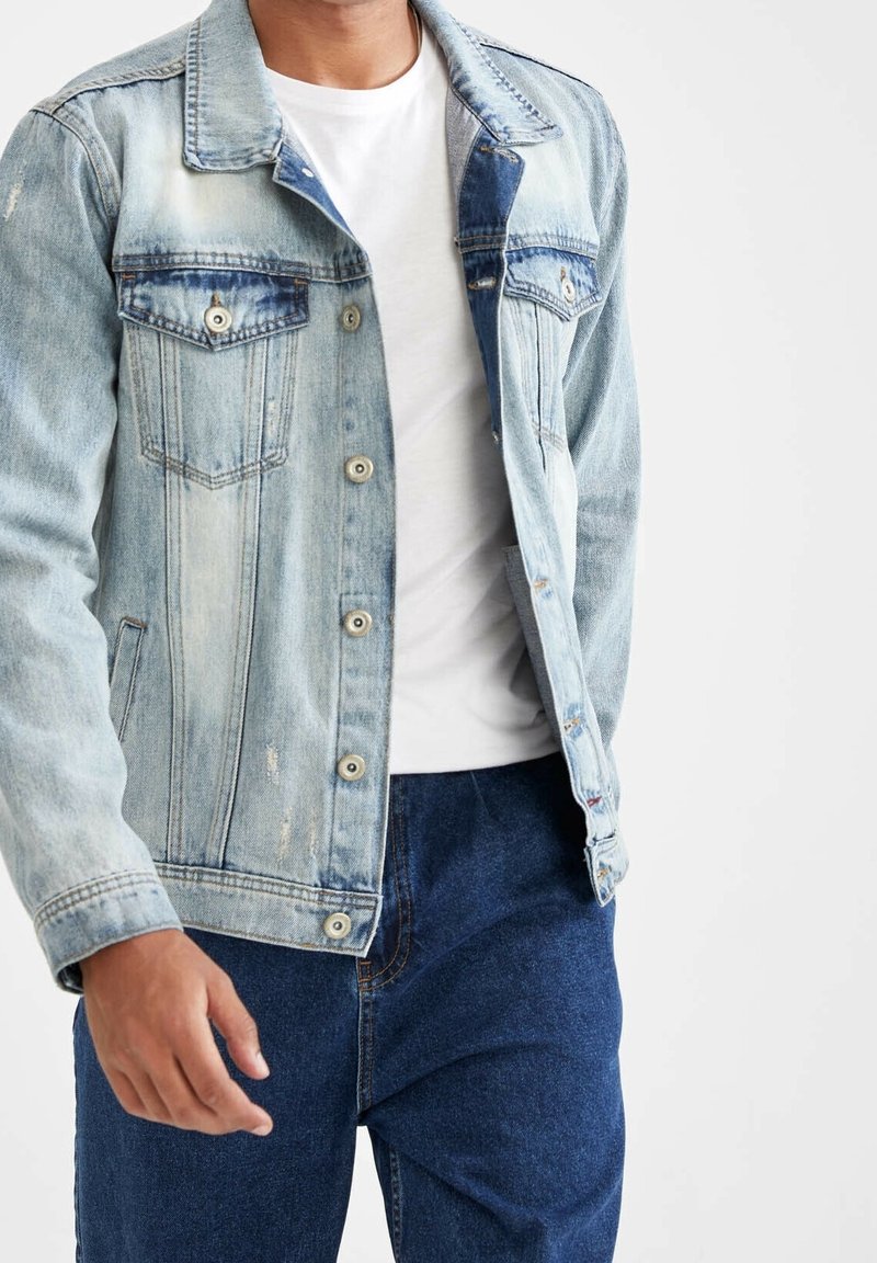 Veste en denim bleu clair avec détails usés, fermetures à boutons métalliques et deux poches poitrine, portée sur un t-shirt blanc et un jean bleu.