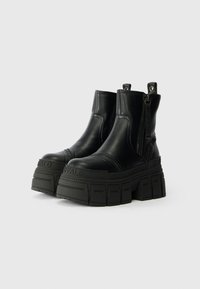 Botas de tornozelo pretas com plataforma, solas chunky, fechos laterais e alças de puxar, exibidas sobre um fundo branco.