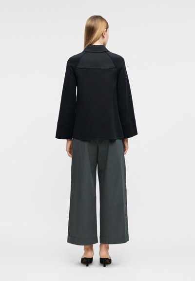 Γυναίκα με ίσια ξανθά μαλλιά φοράει μαύρο μακρυμάνικο σακάκι, γκρι παντελόνι με φαρδιά μπατζάκια και μαύρα παπούτσια με τακούνι, στέκεται με την πλάτη στραμμένη.