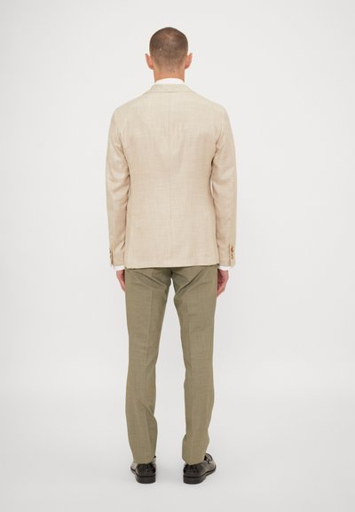 Blazer in lino beige con una vestibilità sartoriale, due spacchi posteriori e pantaloni verde chiaro in un tessuto testurizzato. Le scarpe nere completano l'outfit.