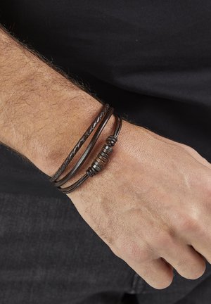Männliches Handgelenk trägt ein mehrsträngiges braunes Lederarmband mit Metallperlen vor einem schwarzen Hemd und dunkler Hose.