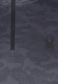 Tissu camouflage gris foncé avec une fermeture éclair noire et un petit logo araignée noir sur le côté droit de la poitrine.