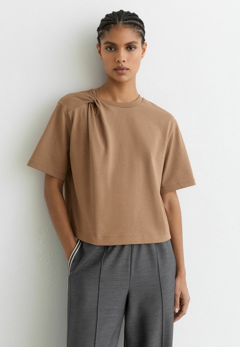 Camiseta marrón de manga corta con un detalle de nudo en el hombro, confeccionada en tela suave; combinada con pantalones grises de corte afilado, que destacan una silueta relajada.