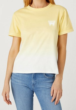 Kvinde iført en kortærmet gul til hvid ombré T-shirt med et lille Wrangler-logo på brystet, kombineret med blå jeans.