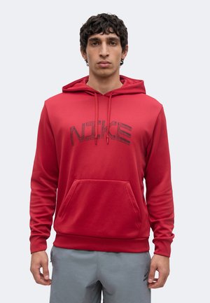 Mladý muž nosí červenú mikinu Nike s kapucňou, predným vreckom a šnúrami, v kombinácii so sivými Nike nohavicami na jednoduchom svetlom pozadí.