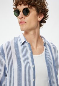 Blauw-wit gestreepte button-up shirt met een kraag, gedragen over een witte tanktop. Ronde groene zonnebril met gouden accenten. Krullend haar.