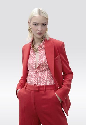 Blazer rojo y pantalones a juego sobre una camisa rosa con estampado. Collar de cadena dorada y pendientes completan el look, que presenta texturas suaves.