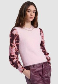 Maglione smanicato rosa chiaro realizzato in materiale morbido, abbinato a una camicia a maniche lunghe bordeaux scuro con motivo floreale. Presenta una scollatura rotonda.