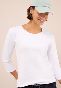Joven sonriendo, vistiendo una camiseta blanca de manga larga, una gorra de béisbol verde claro y pantalones oscuros con cordón, contra un fondo beige.