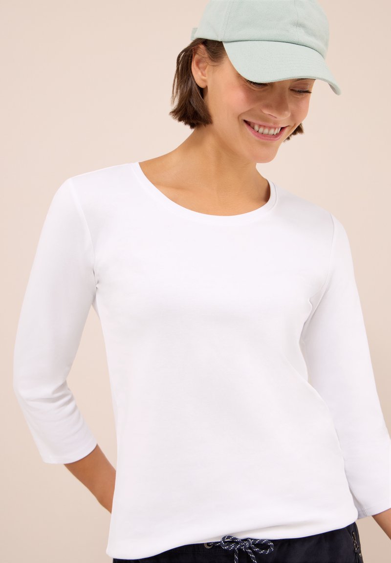 Joven sonriendo, vistiendo una camiseta blanca de manga larga, una gorra de béisbol verde claro y pantalones oscuros con cordón, contra un fondo beige.