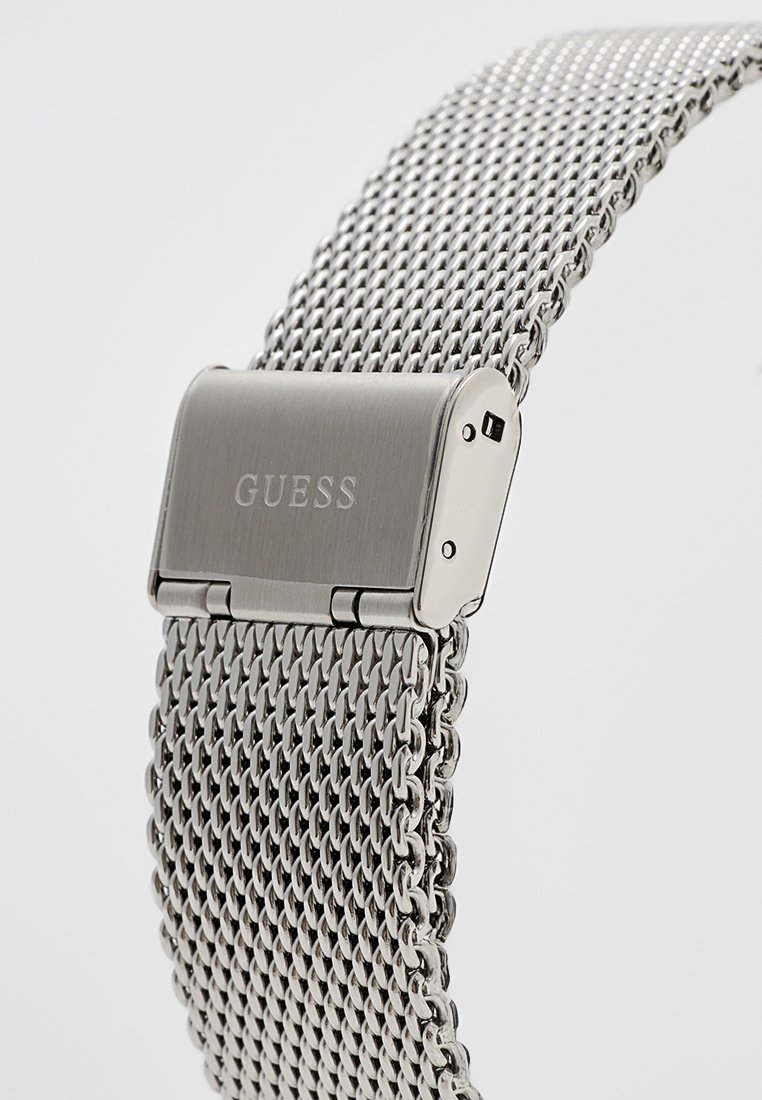 Bracelet de montre en maille argentée en acier inoxydable avec une boucle polie affichant le logo "GUESS". Il présente un design élégant et tissé.
