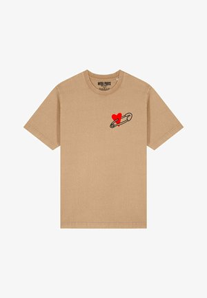 La t-shirt in cotone beige presenta maniche corte, un col rotondo e una grafica di una spilla di sicurezza nera con un cuore rosso sul lato sinistro del petto.