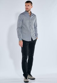 Hemd mit Knopfleiste aus gemustertem blau-weißen Stoff, getragen mit schwarzen Hosen und grauen Sneakers. Bemerkenswerte Details sind der Kragen und die langen Ärmel.