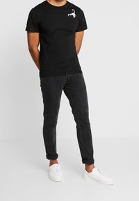 Hombre vistiendo una camiseta negra con un gráfico de langosta blanca, jeans oscuros arremangados y zapatillas blancas, de pie contra un fondo liso.