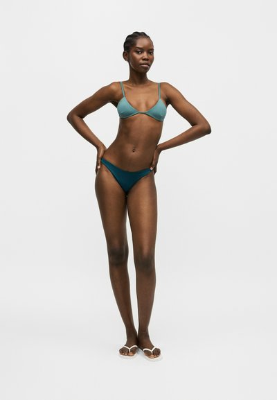 Γυναίκα με ζαφειρί τοπ μπικίνι και σκουρότερη ζαφειρί μπικίνι βερμούδα, στέκεται με τα χέρια στη μέση μπροστά από απλό φωτεινό φόντο.