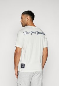 Balts kokvilnas T-krekls ar tumši zilu "New York Yankees" tekstu aizmugurē, īsām piedurknēm un taisnstūra formas logo uzšuvumu apakšmalā.
