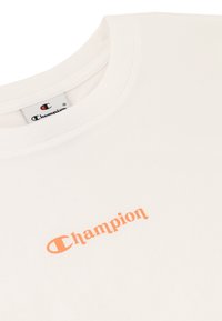 Cremefarbiges T-Shirt mit kurzen Ärmeln, das ein orangefarbenes "Champion"-Logo auf der Vorderseite zeigt. Hergestellt aus weicher Baumwolle mit einem klassischen Rundhalsausschnitt.