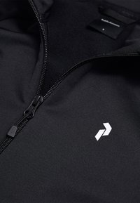 Giacca nera con zip caratterizzata da una texture liscia, un design minimale e un logo bianco sulla parte anteriore vicino alla zip.