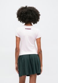 Różowa bluzka z krótkim rękawem o dopasowanym kroju i małym burgundowym logo z tyłu, w połączeniu z ciemnozieloną spódnicą plisowaną.