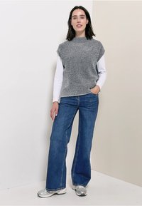 Donna in piedi con una mano in tasca, indossa un maglione grigio senza maniche sopra una camicia bianca a maniche lunghe, jeans blu e sneakers argentate.