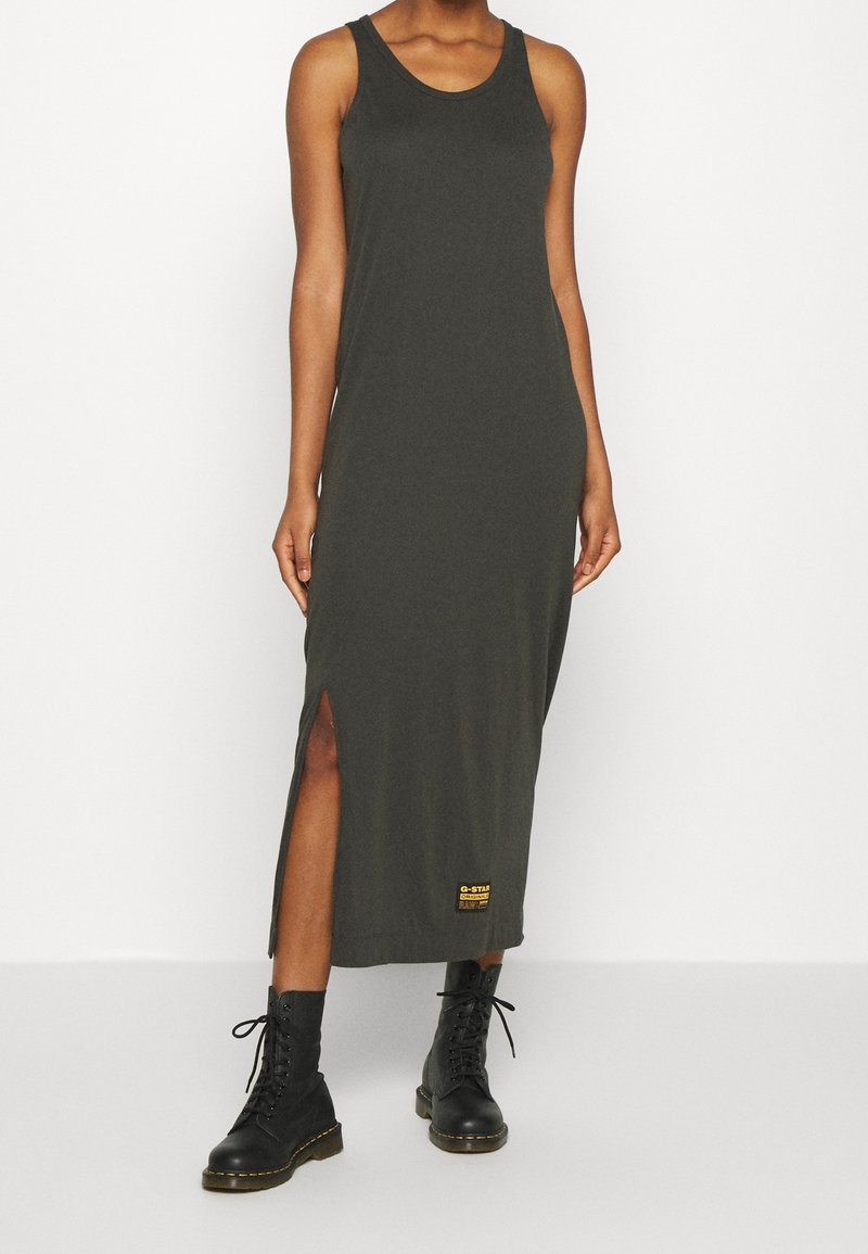 Femme portant une robe maxi sans manches gris foncé avec une fente latérale et des bottes noires à lacets, debout devant un fond blanc uni.